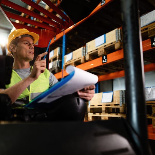 pensive-forklift-operator-examining-stock-packages-shelves-warehouse